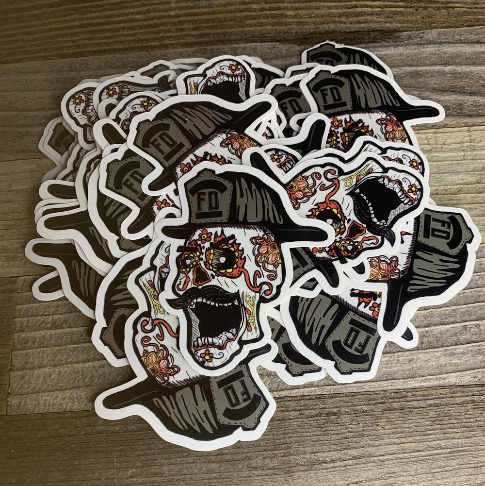 A pile of die-cut stickers featuring a black and white sugar skull wearing a firefighter helmet, decorated with orange and red floral details, displayed on a wooden surface.