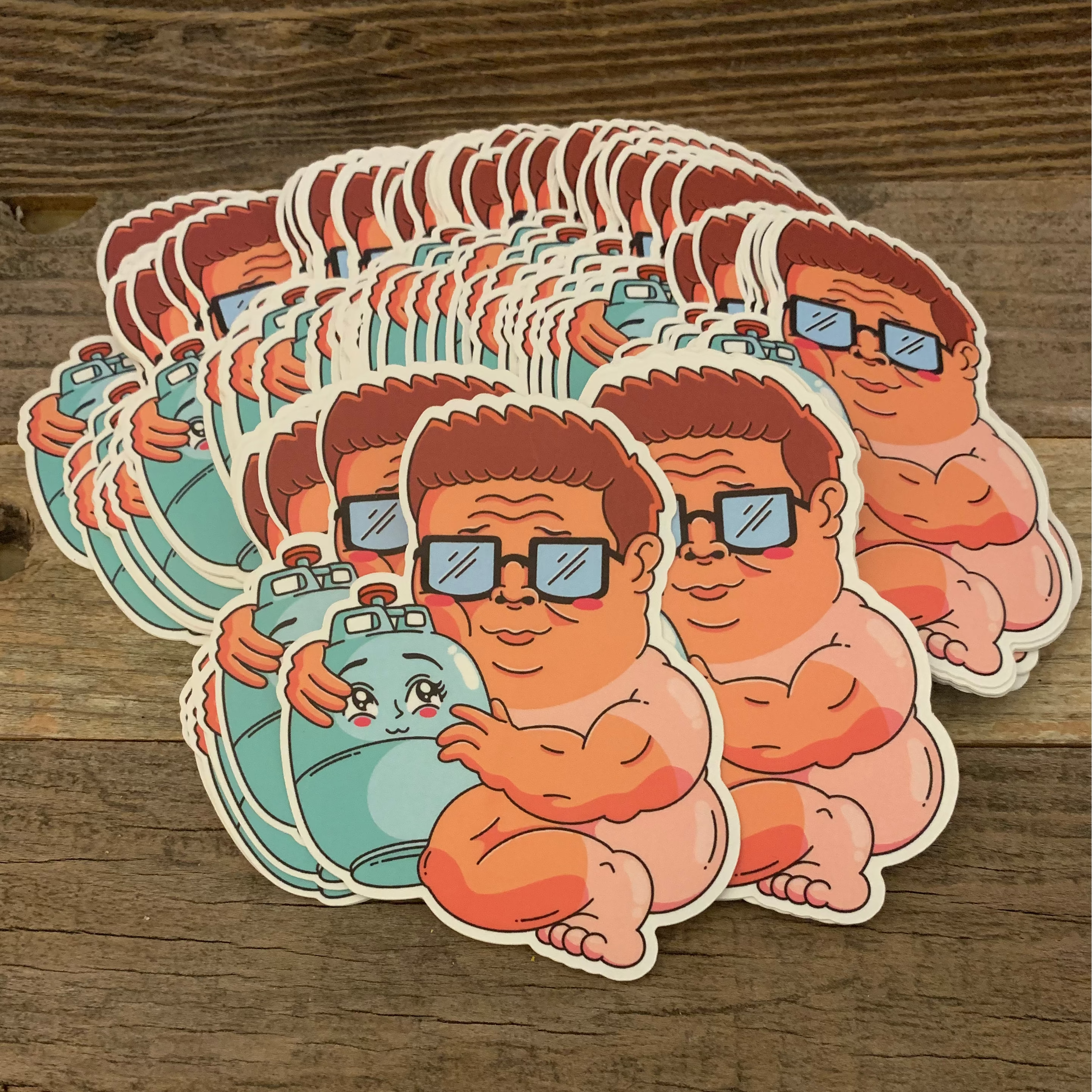 A stack of cartoon die-cut stickers featuring a baby Hank Hill character with brown hair, blue sunglasses, and a blue gas tank, displayed on a rustic wooden surface.
