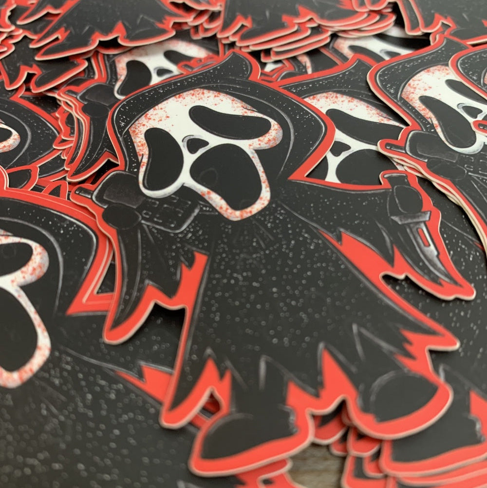 A close-up of multiple Ghostface-shaped stickers arranged on a dark surface. The stickers are black with a red outline and a white screaming face graphic.